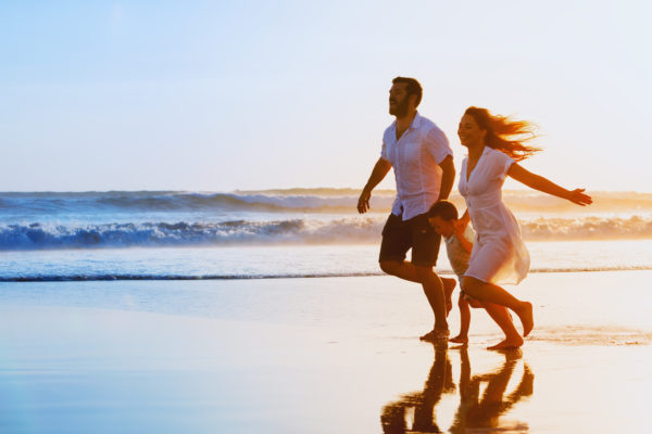 Happy family – father, mother, baby son hold hands and run together with fun along sunset sea surf on black sand beach. Travel, active lifestyle, parents with children on tropical summer vacations. Fitness Holidays and Retreats